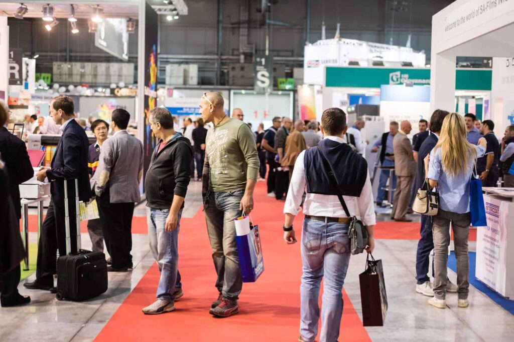 Attendees walking through a busy trade show exhibition hall with booths on both sides, networking, browsing displays, and carrying branded bags along a red carpet aisle.