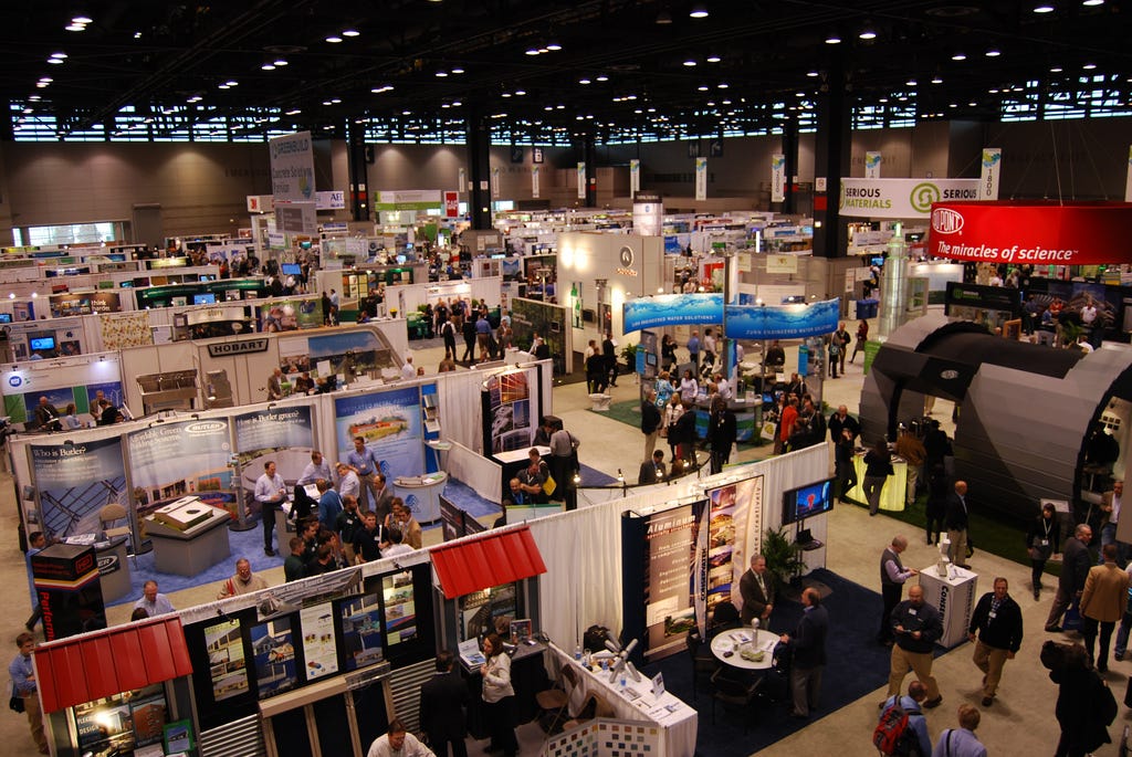 Wide view of a busy indoor trade show floor with multiple exhibition booths, branded displays, and attendees networking inside a large convention hall.