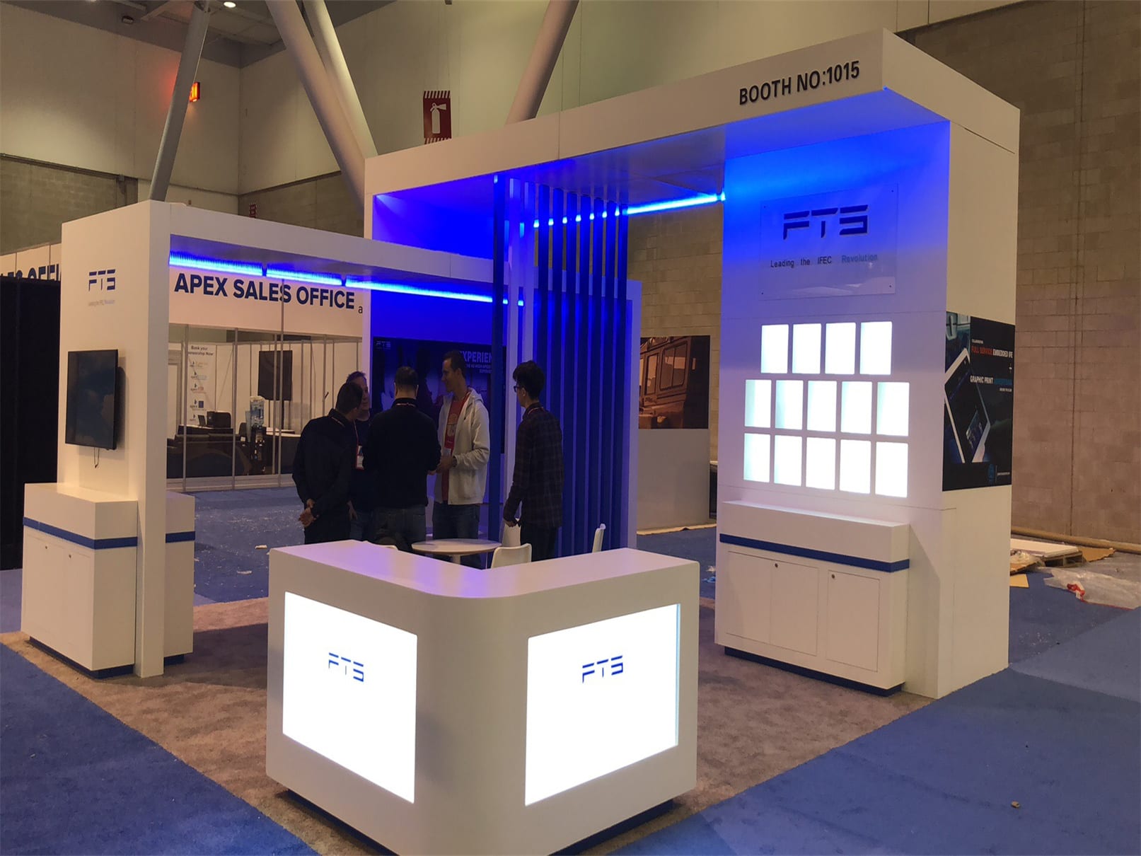 Modern custom trade show exhibit booth with white modular structures, blue LED lighting, illuminated counters, digital display panels, and open meeting space inside an expo hall.