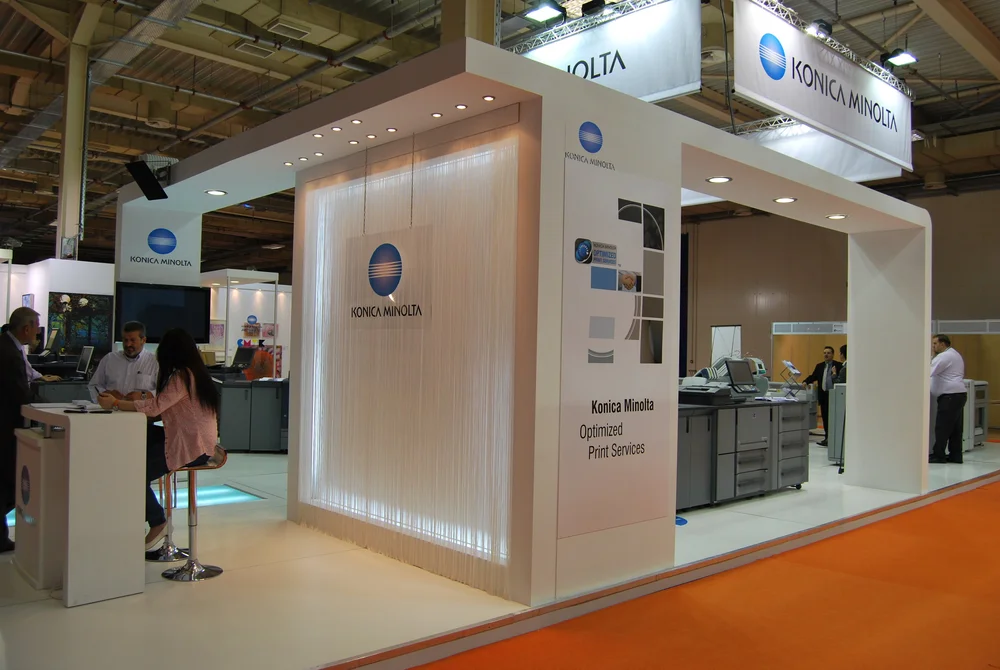 Modern Konica Minolta trade show booth with white modular structure, branded signage, demo counters, and staff engaging visitors inside an exhibition hall.