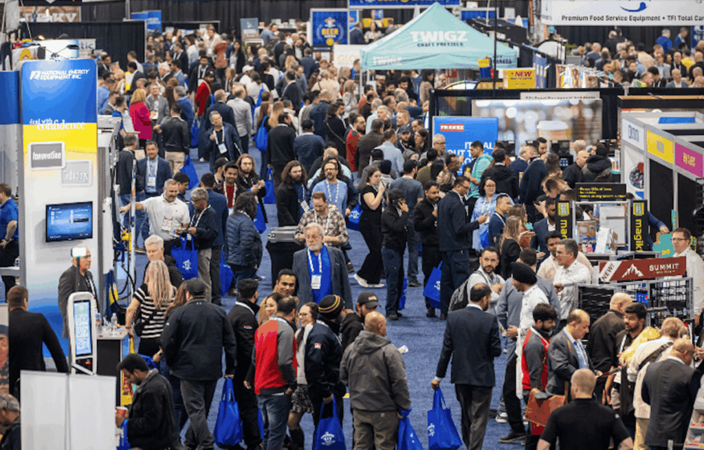 Crowded trade show floor with busy exhibition booths, large crowds of attendees, branded displays, and professionals networking in a convention hall.